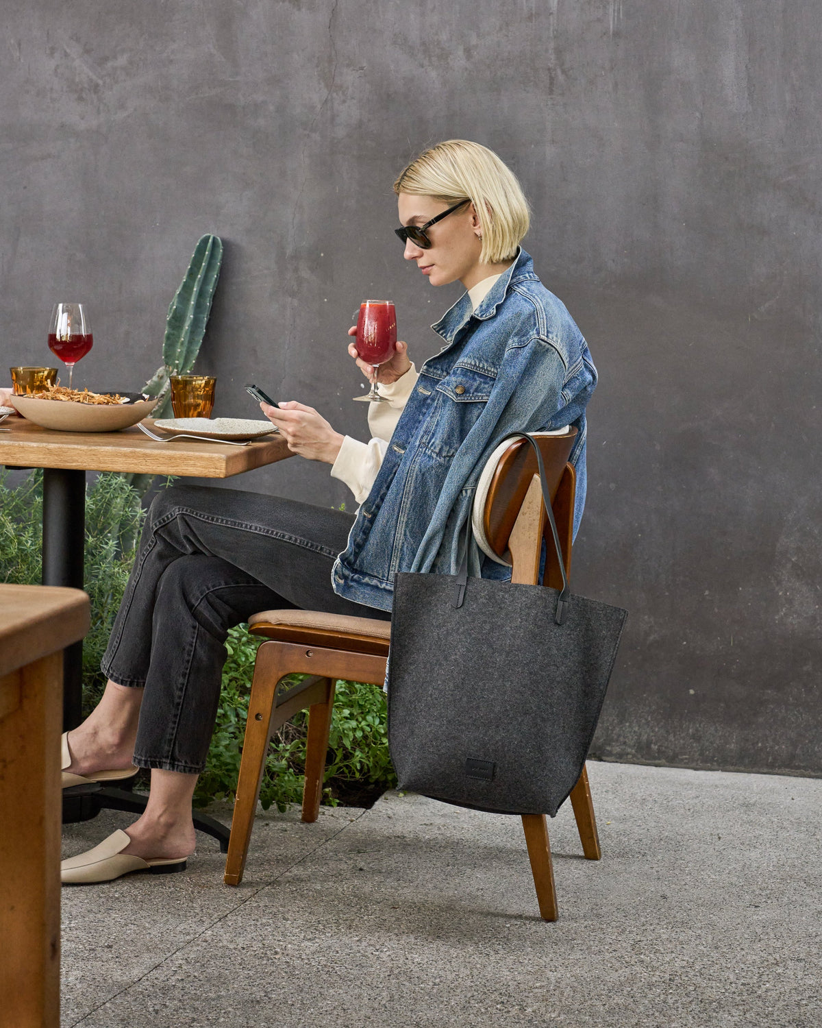 Chic woman sitting in a restaurant with the leather straps of a Hana Merino Wool Felt Tote elegantly hanging over the chair back