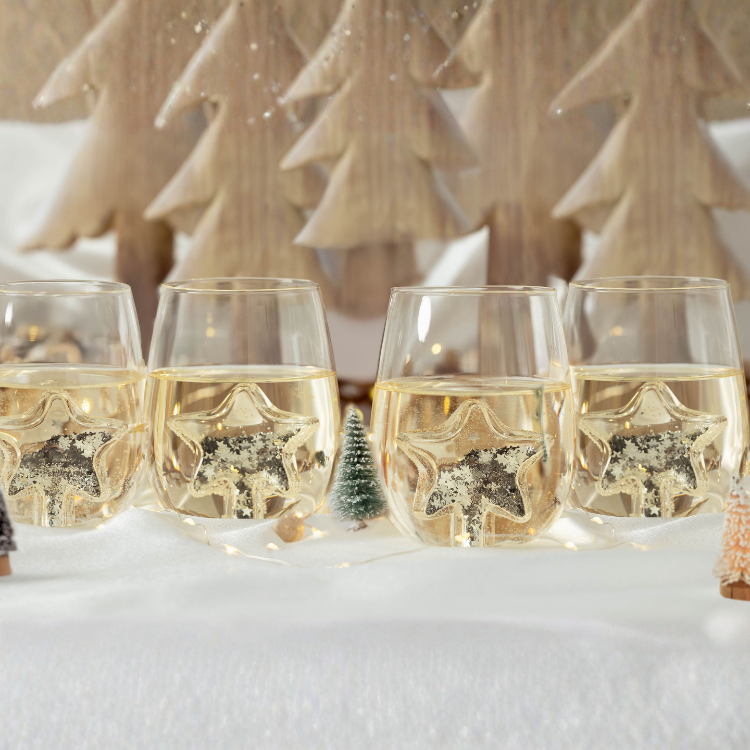 Four glasses of champagne with decorative stars in a snowy scene with Christmas trees.