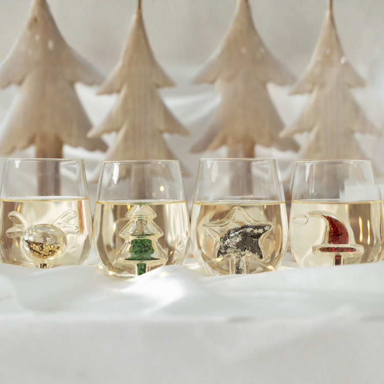 Four stemless wine glasses with Christmas-themed decorations in a snowy setting.
