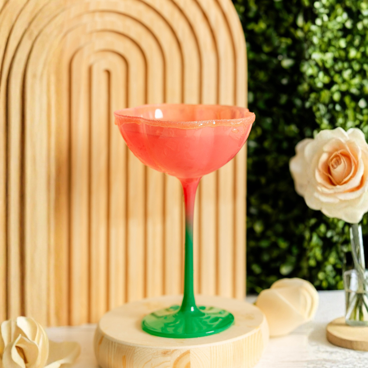 Colorful glass with a green base and pink top on a wooden stand, with a blurred background of greenery and flowers.