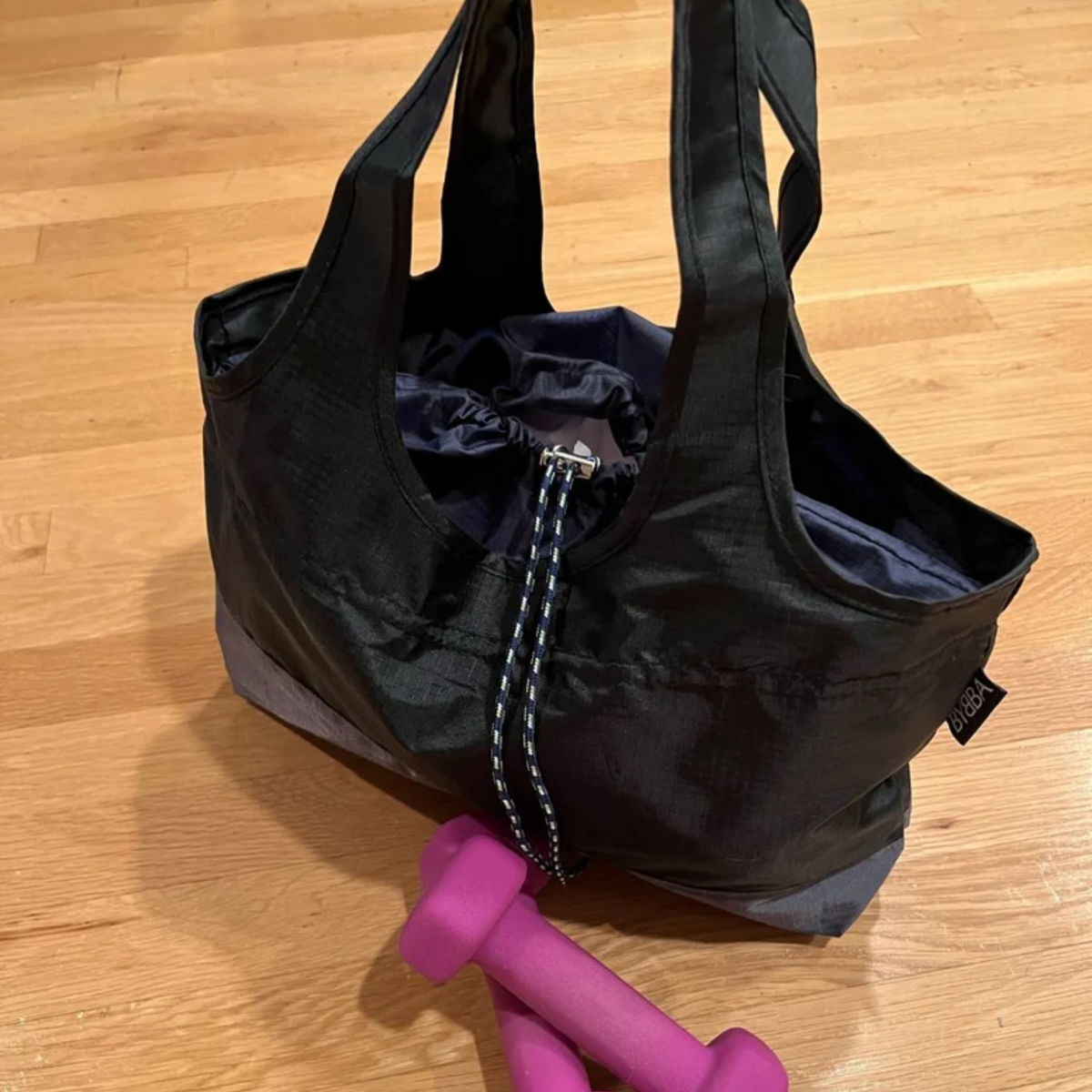 Black packable, foldable, reusable tote made from recycled ocean plastic by BYBBA, shown with pink dumbbells on wood floor.