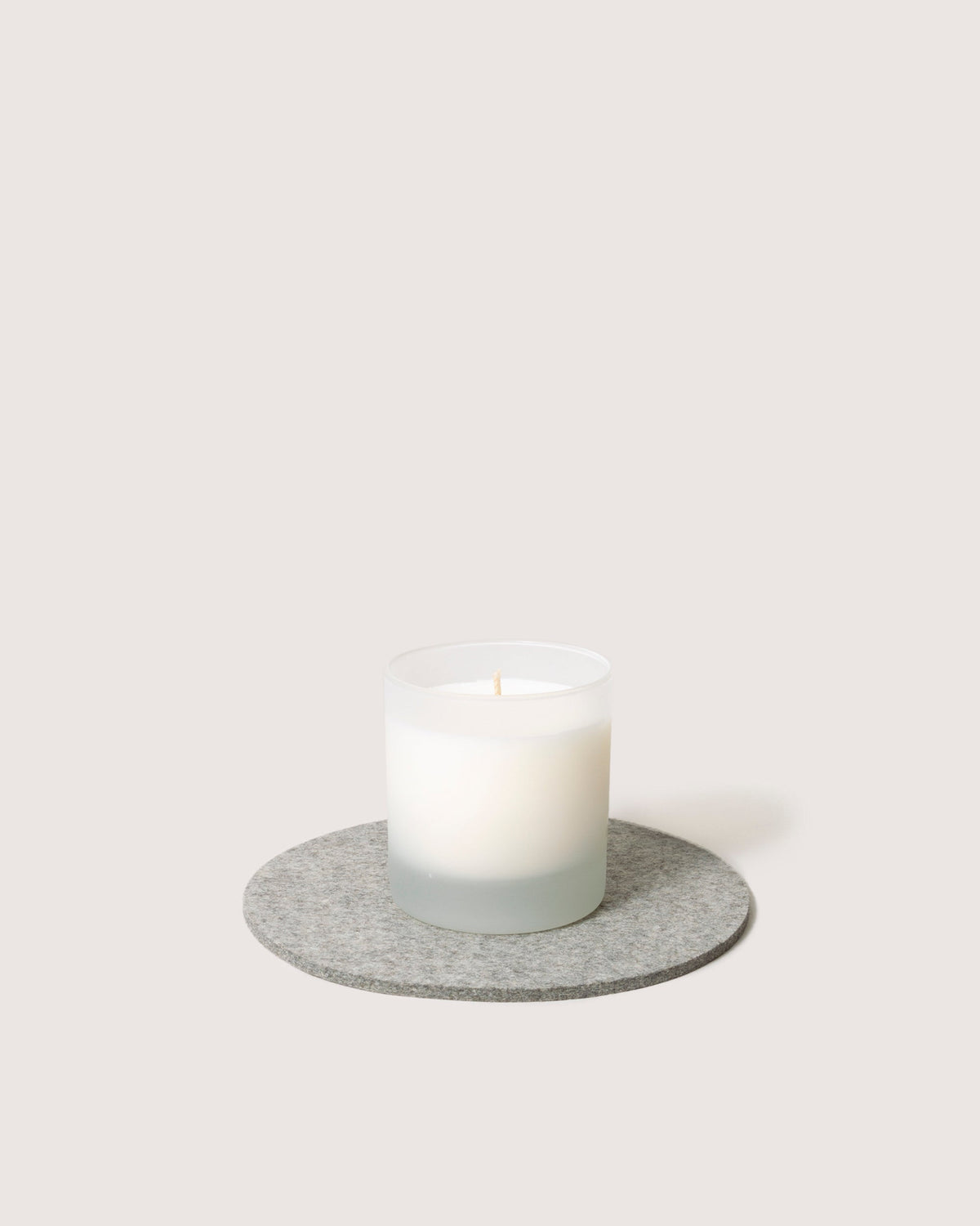 A lit white candle in a frosted glass holder sits on a round gray felt mat made of merino wool by graf lantz, set against a plain light background.