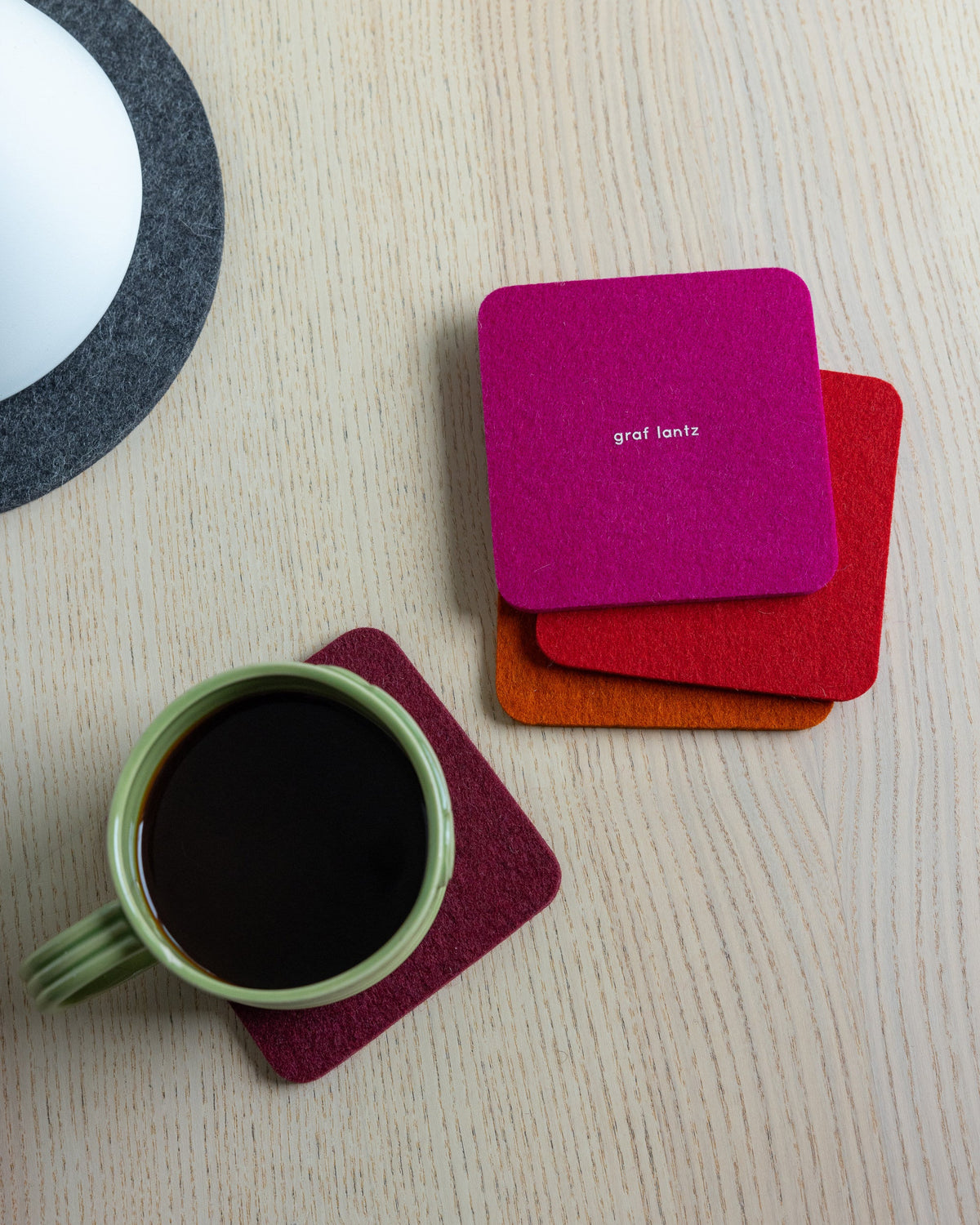 A green mug of black coffee sits on a magenta graf lantz coaster crafted from German Merino wool, atop a light wooden table. Three coasters in magenta, orange, and red are artfully arranged nearby. A hint of a white and gray object is visible at the edge.