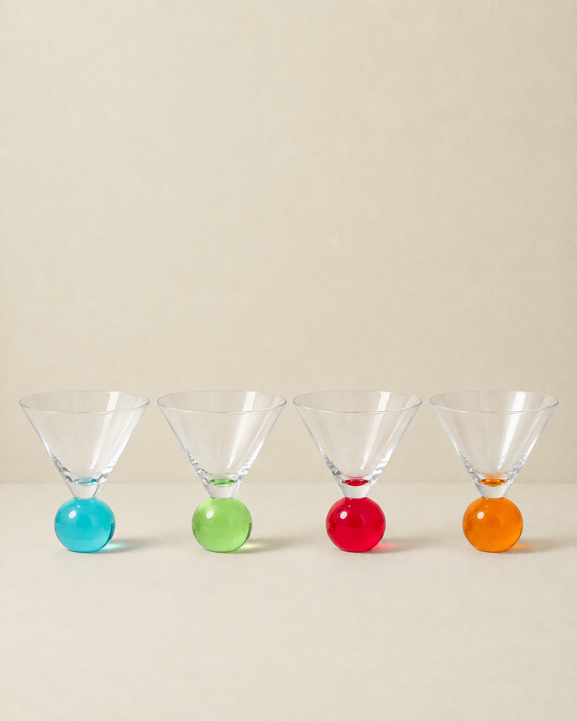 Four martini-style glasses with clear conical bowls and colorful spherical bases in blue, green, red, and orange, arranged in a row against a warm beige background.