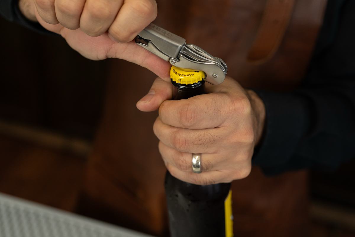 Close-up of hands using a silver professional wine opener to pop open a beer bottle.