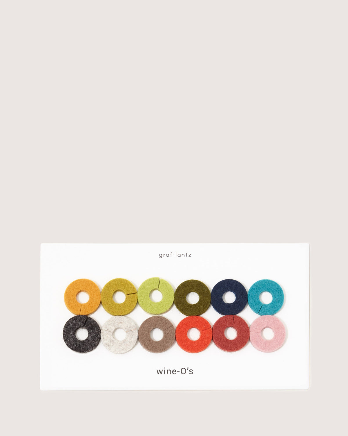 The graf lantz felt wine glass markers offer a sustainable luxury touch with circular designs in vibrant colors like orange, yellow, green, blue, gray, and pink. Presented on a white card with graf lantz and wine-Os labeling for a chic color code.