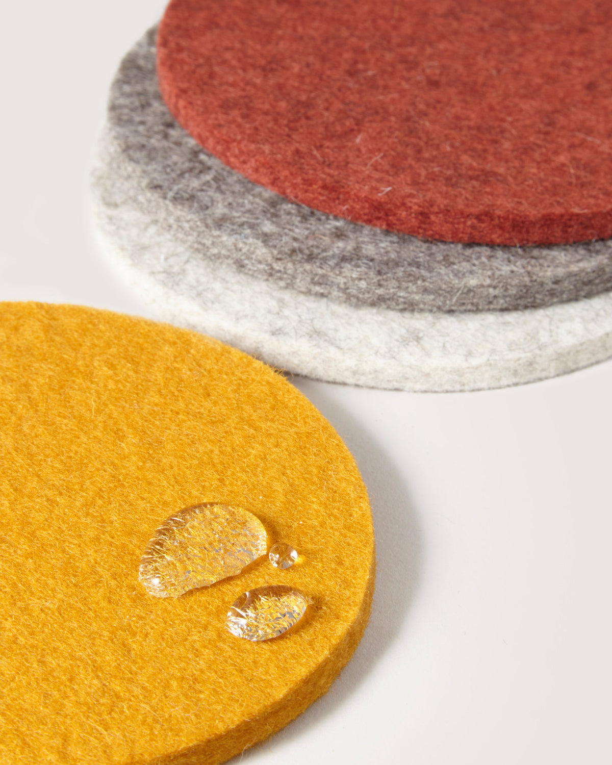A set of graf lantz round Merino wool coasters in yellow, white, gray, and red is showcased. The foreground features a yellow coaster with water droplets, highlighting its water resistance. Perfect as beer felt coasters, they are placed on a plain light-colored surface.