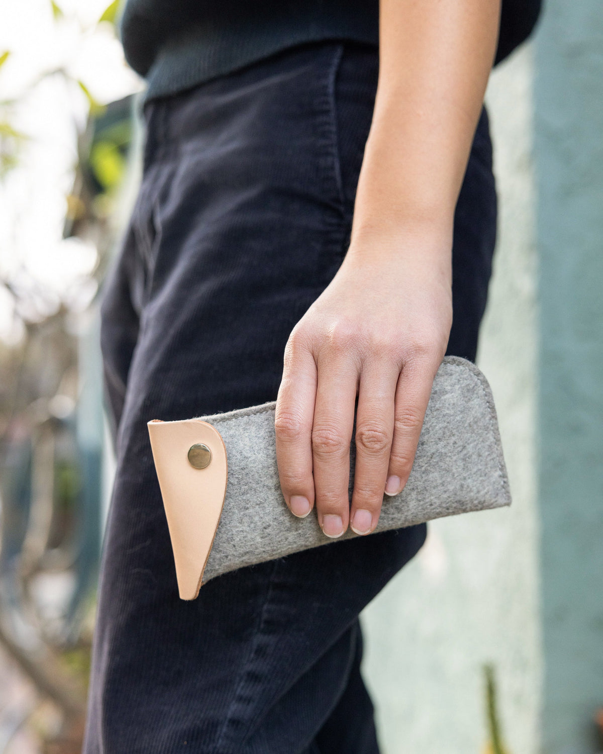 A person holds a chic graf lantz gray felt eyeglass sleeve with a tan leather flap against a softly blurred outdoor backdrop of lush greenery and hints of a turquoise wall, exuding an effortlessly stylish vibe in dark pants and top.