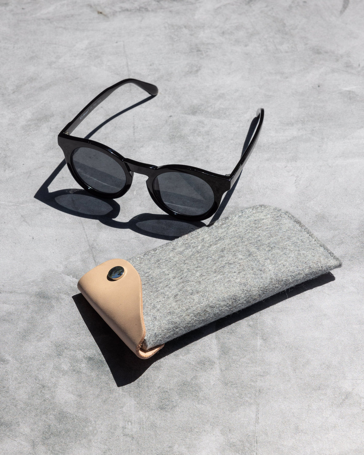 Black round sunglasses sit on a flat gray surface beside a graf lantz eyeglass sleeve, crafted from gray felt with a tan leather flap and button closure.