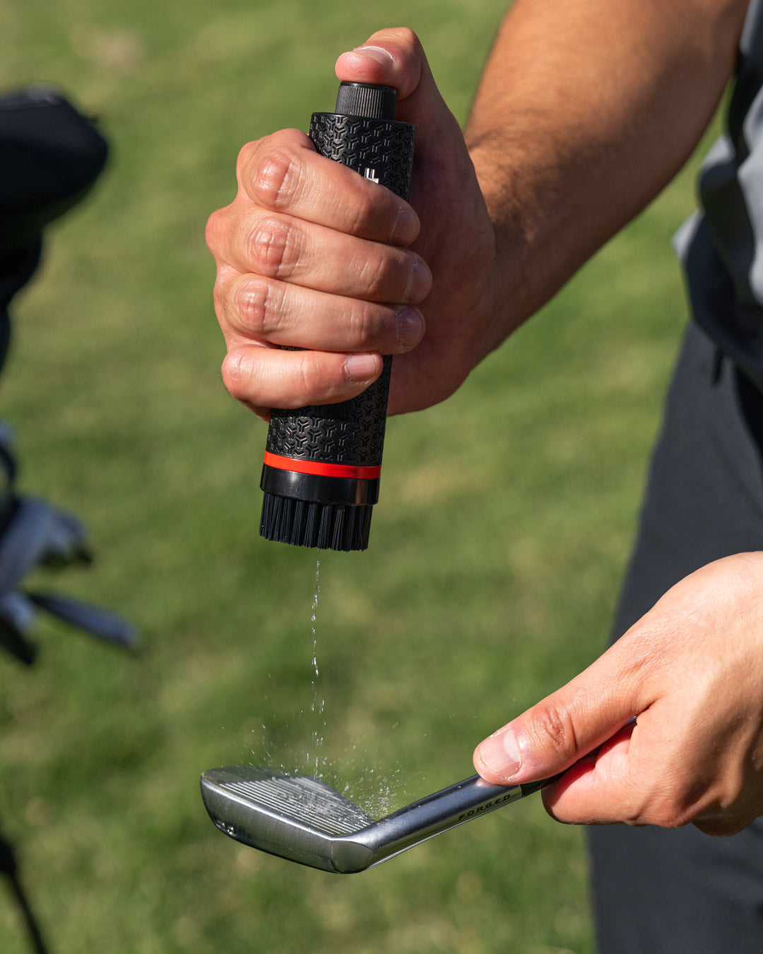 Person applying a water to a golf club head using a handheld Golf Brush Cleaner on a grassy background. #color_navy