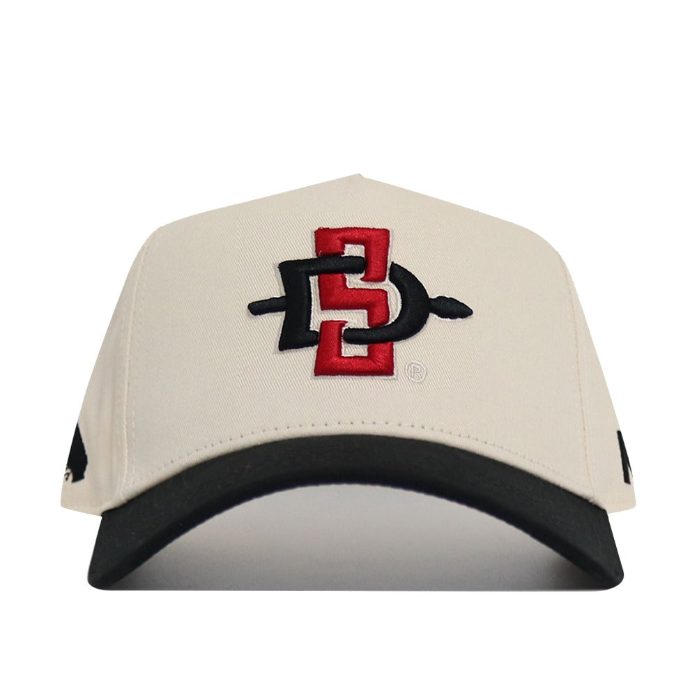 The No Rivals Classic San Diego State Hat (B2B) is a beige snapback with a black brim and red & black SDSU interlocking logo embroidered on the front, crafted from high-quality cotton for a comfortable, structured fit.