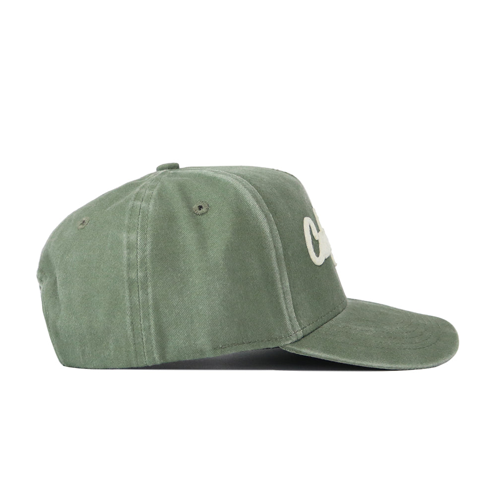 The California Home Turf Hat is shown in a side view. Made from washed cotton, this green snapback features ventilation eyelets and a curved brim, with white embroidered text partially visible on the front panel.