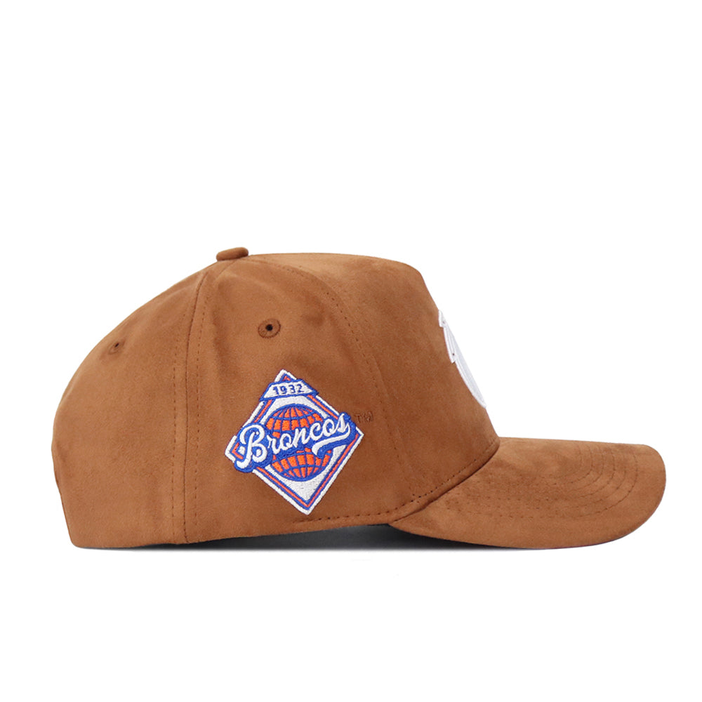 Boise State Heritage Hat by No Rivals, shown from the side, features a blue and white embroidered patch on the left panel. Made with suede fabric, it has a curved brim, visible stitching, and an adjustable snap back closure.