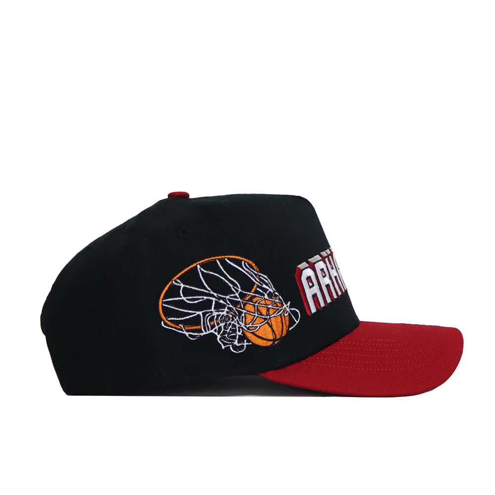 The Arkansas Courtside Hat is a black and red snapback with a flat brim, white block text on the front, an embroidered abstract basketball and net design on the side, and a structured fit made from high quality cotton.