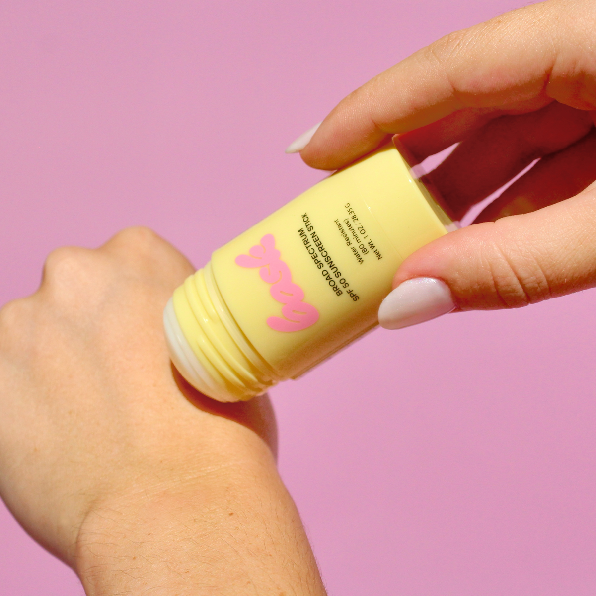 Hand applying a yellow stick of sunscreen with a pink logo on a pink background