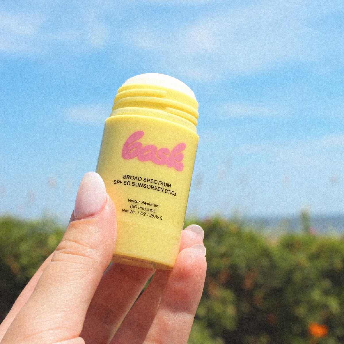 Hand holding a yellow sunscreen stick with a blue sky and greenery in the background