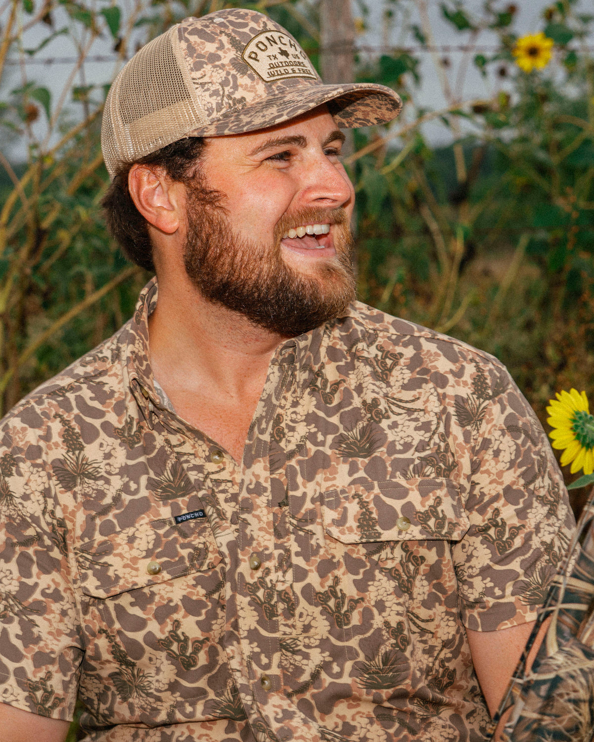 Man smiling outdoors wearing The Albany shirt with a nature-inspired camo pattern and a mesh trucker hat