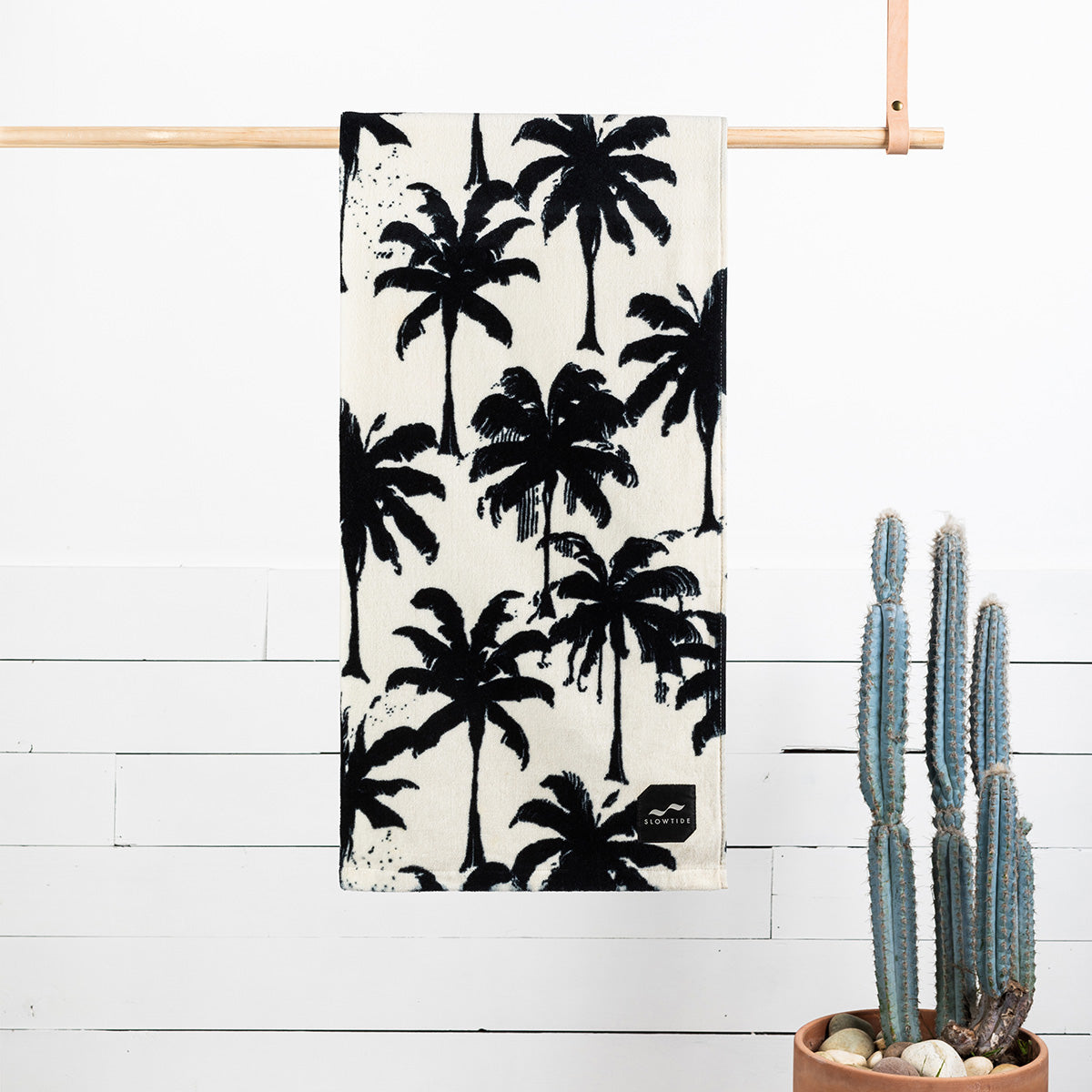Beach towel with black palm tree print displayed beside a cactus against a white wall.