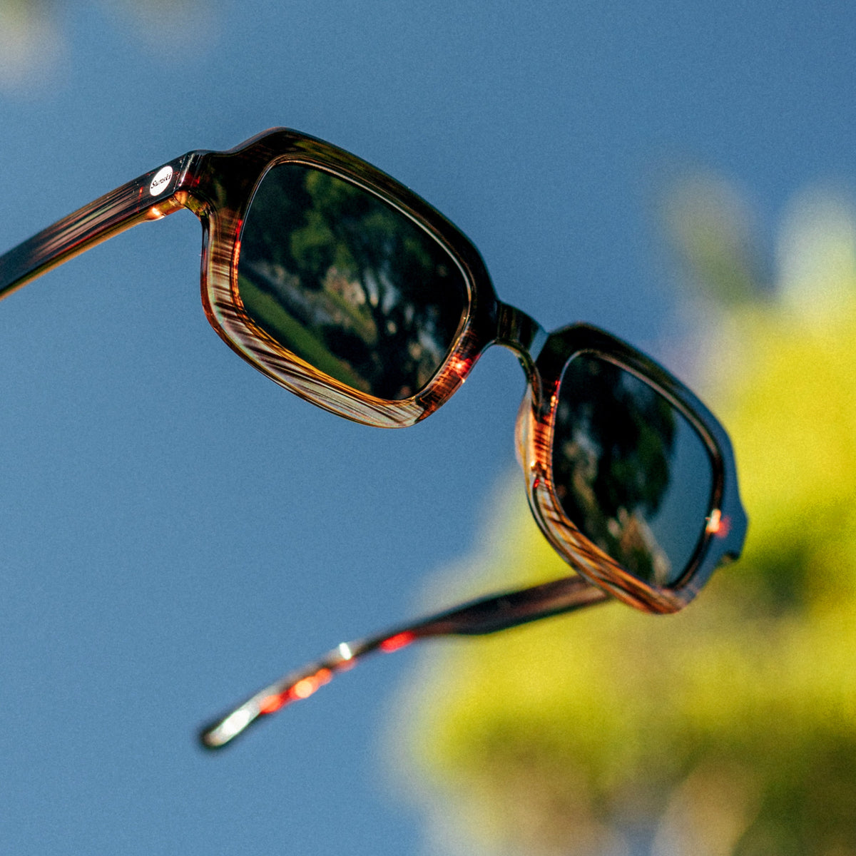 Sunglasses with reflective lenses against a blue sky and blurred greenery