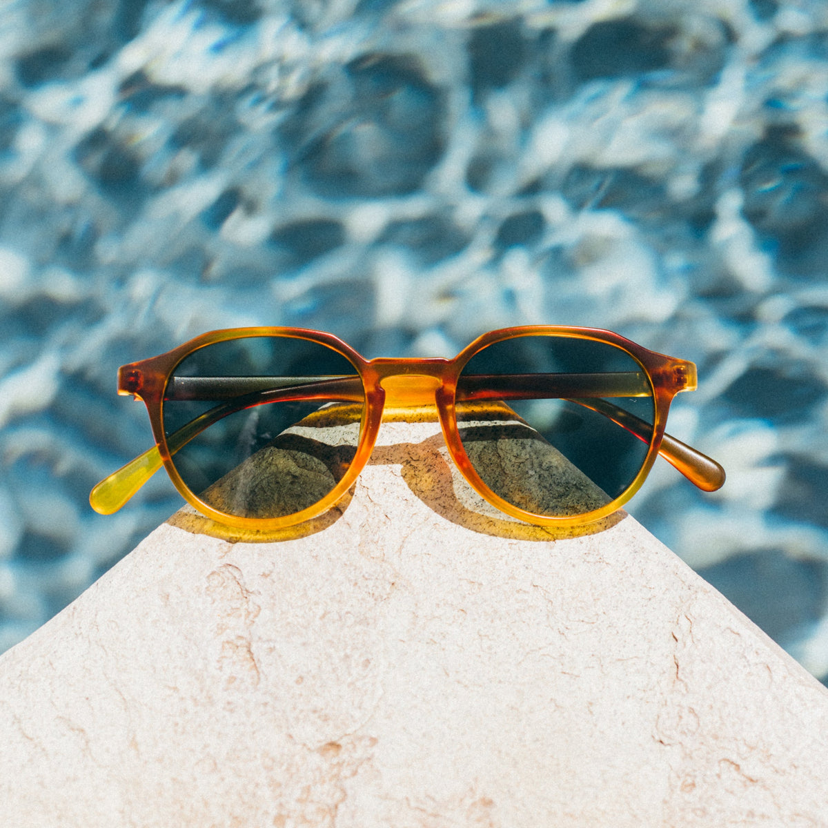 Sunglasses with orange frame and reflective lenses on a rock with water in the background