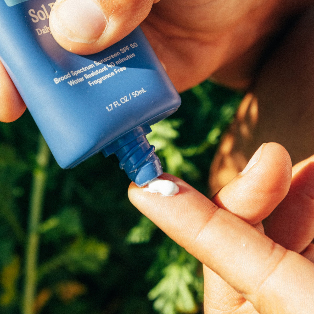 Person applying sunscreen to their hand with a blue bottle against a blurred green background