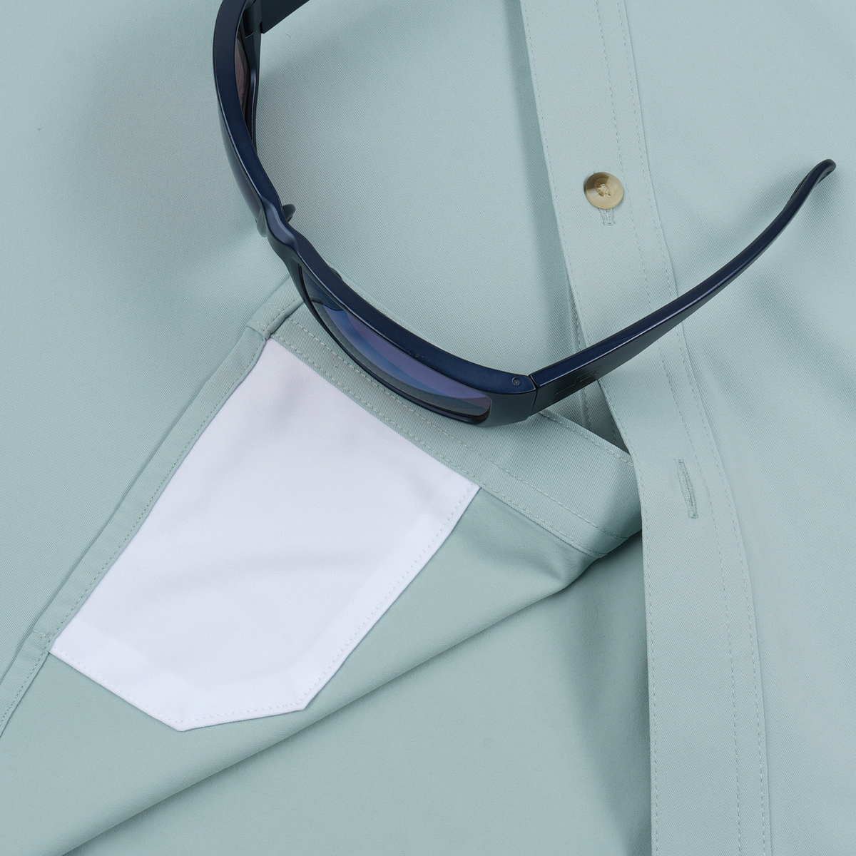 Close-up of a light green shirt with a white pocket and dark blue sunglasses resting on it.