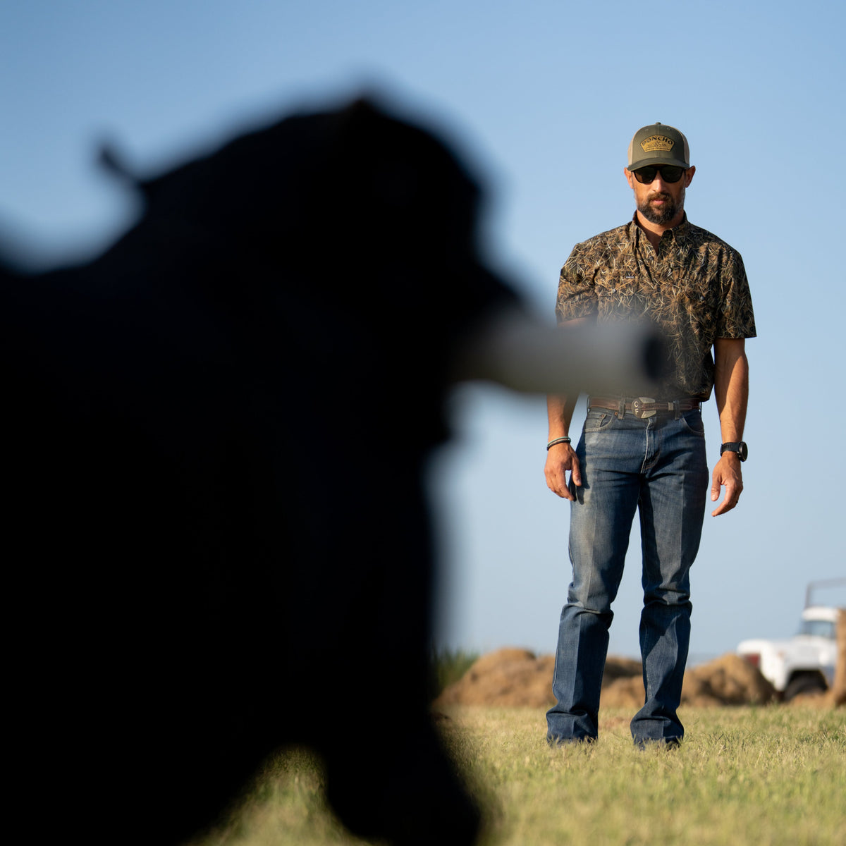 Man wearing The Layout Blind shirt and jeans standing outdoors on grass with a blurred large black animal in the foreground
