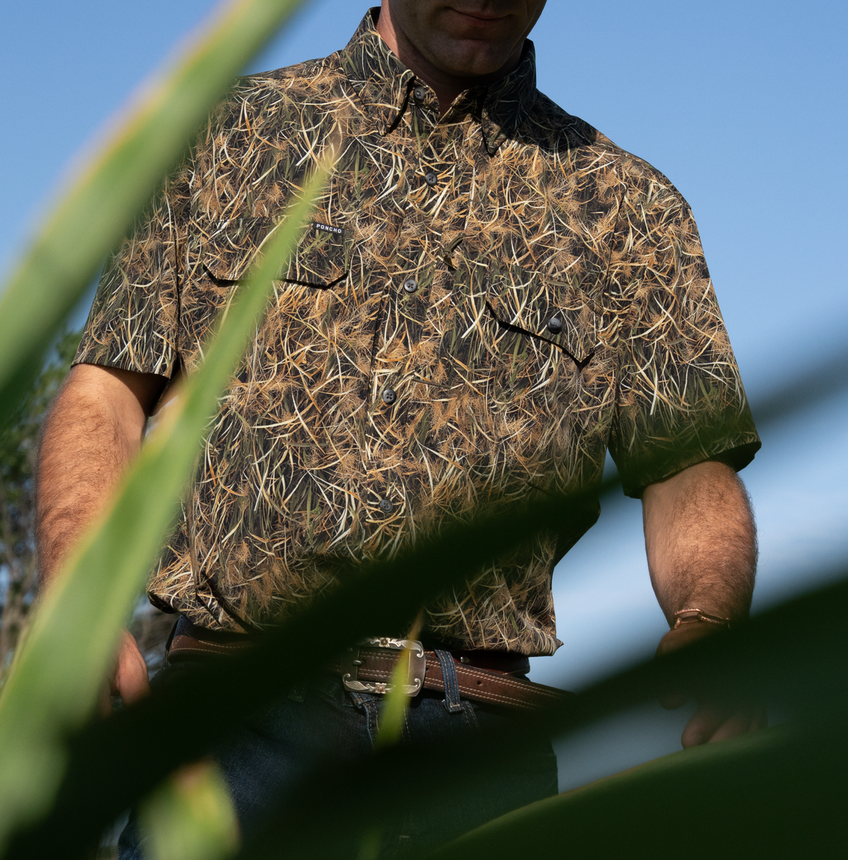 Man wearing The Layout Blind shirt with a nature-inspired camouflage print standing outdoors among tall green plants under a clear blue sky