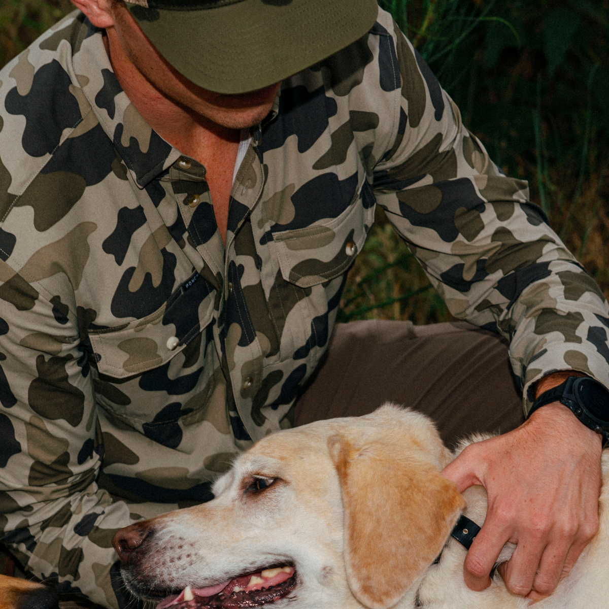 Man wearing The Zenaida camo shirt outdoors petting a yellow Labrador retriever dog