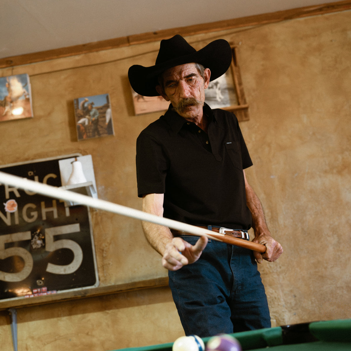 Man wearing black shirt and cowboy hat playing pool indoors with The Permian shirt