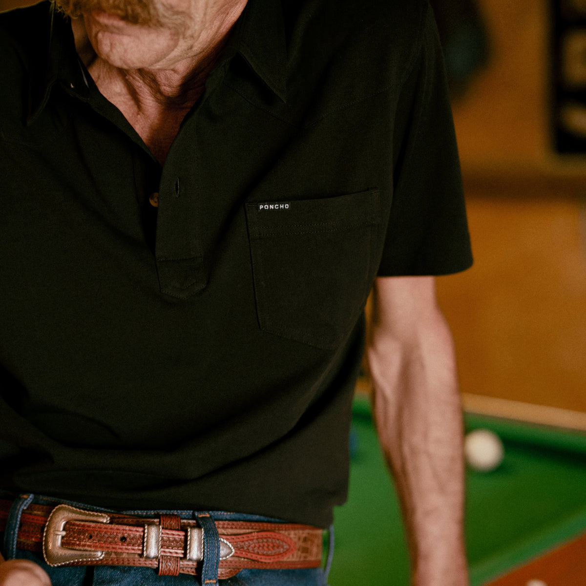Man wearing The Permian black shirt leaning on a pool table with a brown leather belt visible