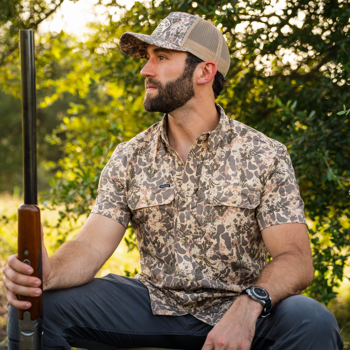Man wearing The Albany shirt with a nature-inspired pattern sitting outdoors holding a shotgun and wearing a mesh back cap