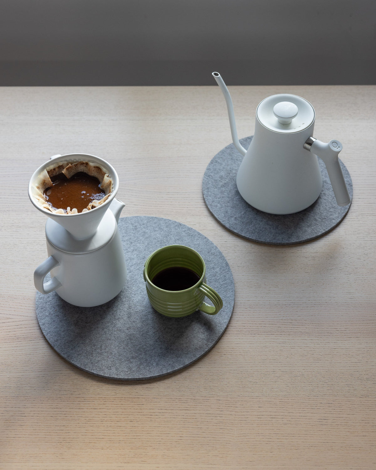 A light wooden table holds a pour-over setup featuring a white ceramic dripper and carafe. A green mug sits on a graf lantz gray moisture-wicking felt mat, while a white gooseneck kettle rests on an identical heat-resistant graf lantz mat.