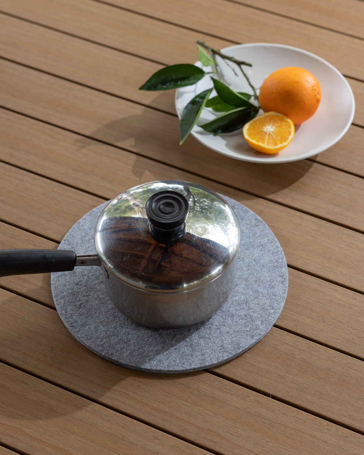 A small metal saucepan with a black handle sits on a graf lantz gray felt mat crafted from moisture-wicking Merino wool, placed on a wooden surface. Beside it is a white plate holding an orange, a halved orange, and some green leaves.