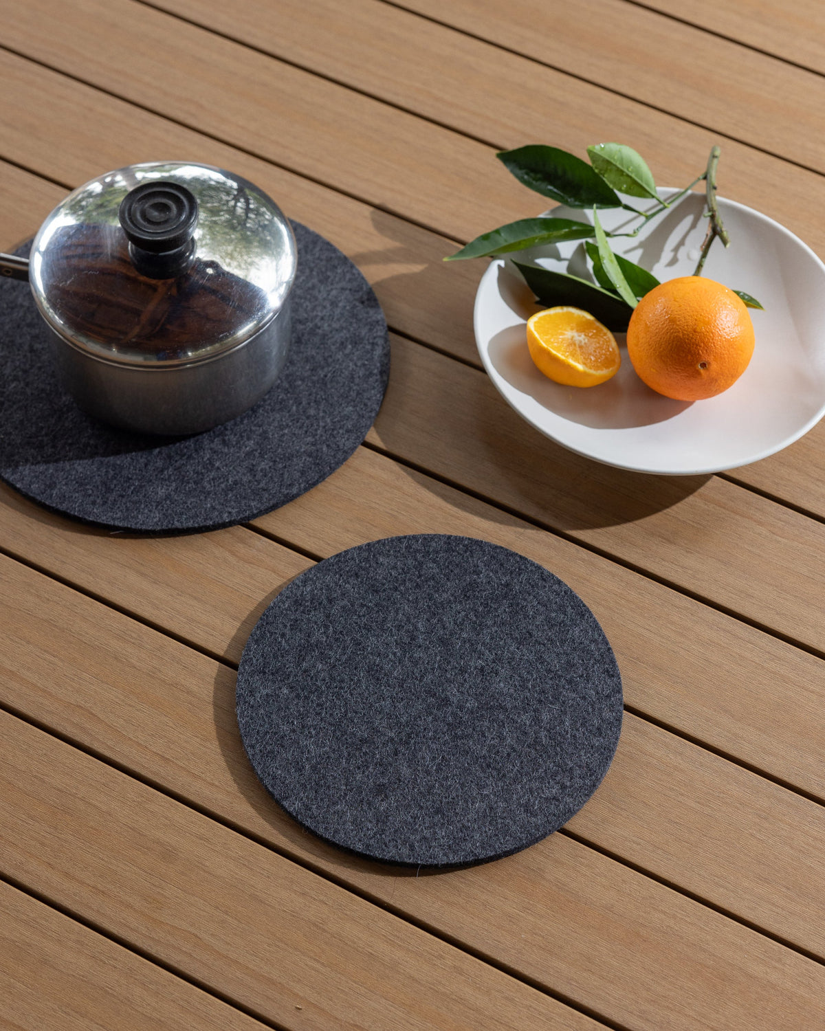 A metal pot with a dark lid rests on a round gray Merino wool mat by graf lantz, highlighting its heat-resistant quality on a wooden surface. Nearby, a white bowl holds an orange, half an orange, and green leaves. An identical trivet is placed in front.