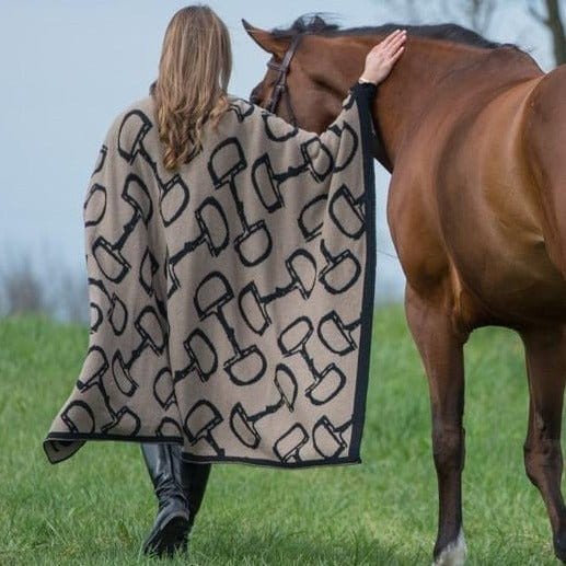 A person wearing a patterned poncho walks beside a brown horse on a grassy field, gently touching the horse’s neck, while the in2green Reversible Bits - Sustainable Knit Equestrian Throw Blanket rests nearby like an equestrian blanket, adding comfort and charm to the country house setting.