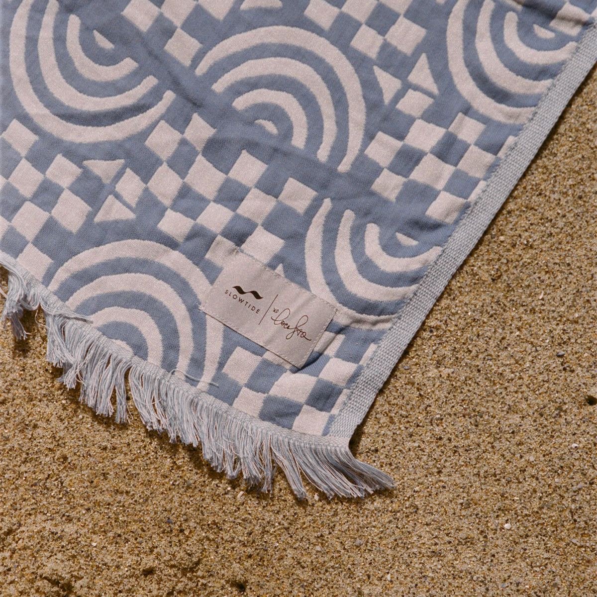 Beach blanket, blue and white woven fabric with fringe, on sand.