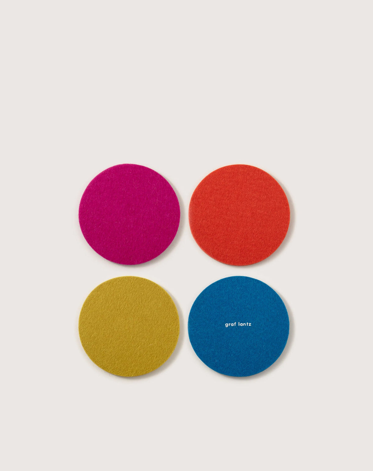 A square arrangement on a white background features four round Merino wool coasters in pink, orange, yellow, and blue. Graf Lantz is printed on the blue one, highlighting Beer Felt craftsmanship quality.