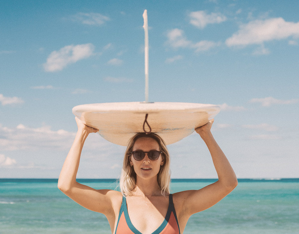 STORY girl holding surfboard over head wearing sunsk yuba sunglasses