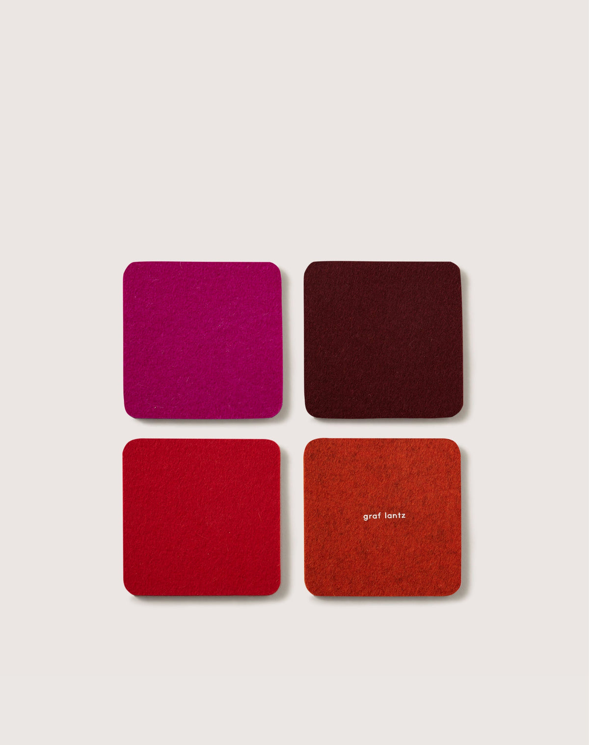 A 2x2 grid of four square coasters from graf lantz, crafted from luxurious German Merino wool. They come in vibrant colors: pink, burgundy, red, and orange. The orange coaster has graf lantz printed in small white letters.