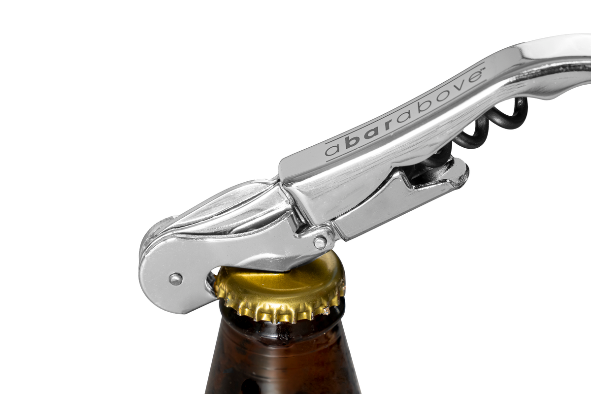 A chrome wine opener with a black corkscrew detail actively opening a brown beer bottle, angled upwards.
